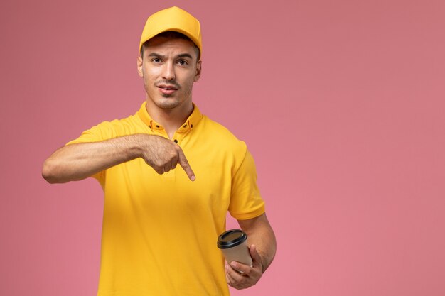 Front view male courier in yellow uniform holding brown coffee delivery cup on the pink background  
