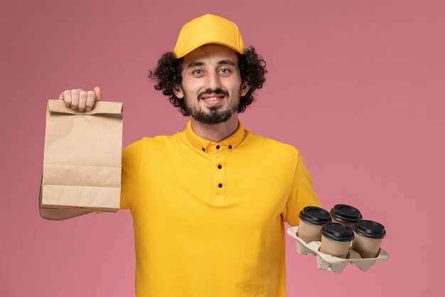 Front view male courier in yellow uniform holding brown delivery coffee cups and food package on the pink wall