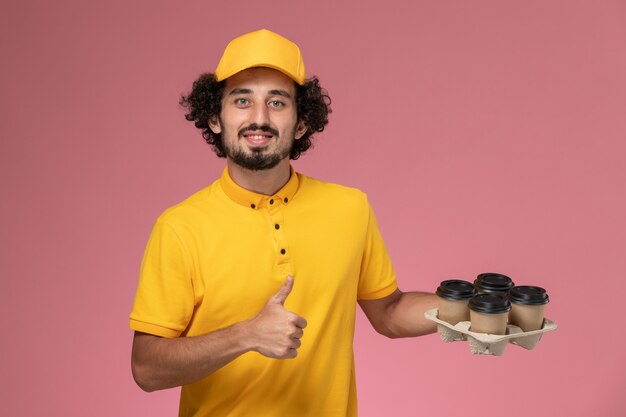 Курьер-мужчина в желтой форме, держащий коричневые кофейные чашки на розовой стене, вид спереди
