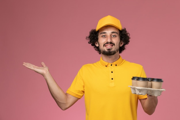 Front view male courier in yellow uniform holding brown delivery coffee cups on pink wall