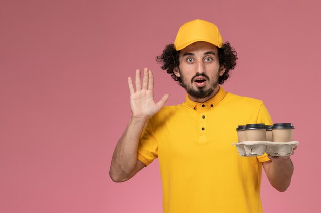 Front view male courier in yellow uniform holding brown delivery coffee cups on pink wall