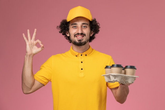 Front view male courier in yellow uniform holding brown delivery coffee cups on the pink wall