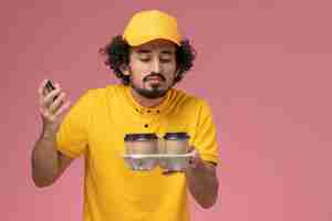 Free photo front view male courier in yellow uniform holding brown delivery coffee cups smelling scent on pink wall