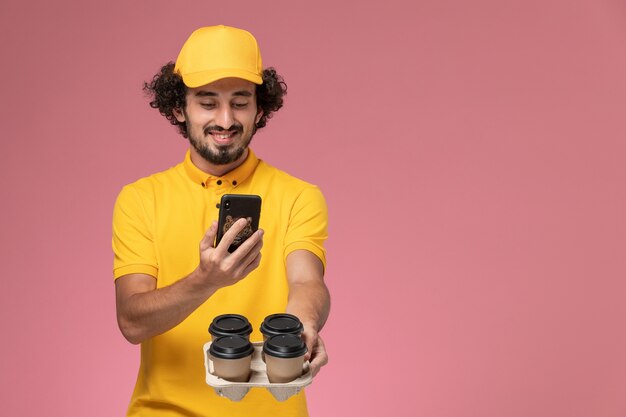 Курьер-мужчина в желтой униформе, вид спереди, держит коричневые кофейные чашки и фотографирует их на розовой стене