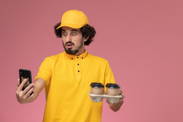 Front view male courier in yellow uniform holding brown delivery coffee cups talking on phone on pink wall