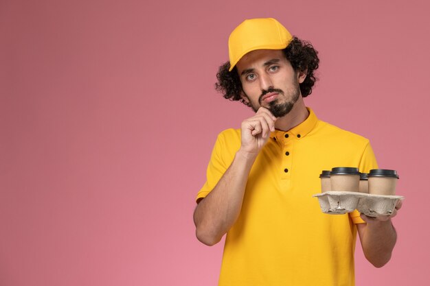 Front view male courier in yellow uniform holding brown delivery coffee cups and thinking on pink wall