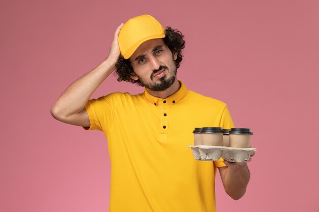 Front view male courier in yellow uniform holding brown delivery coffee cups thinking on pink wall