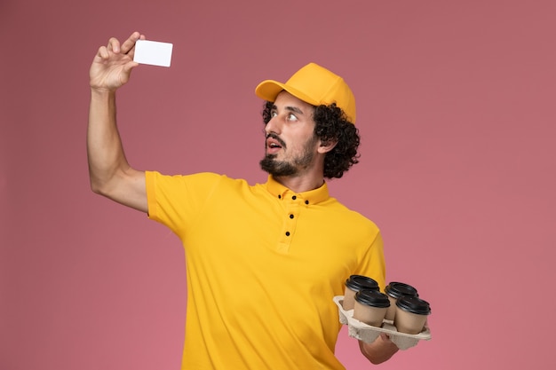 Front view male courier in yellow uniform holding brown delivery coffee cups and white card on the pink wall