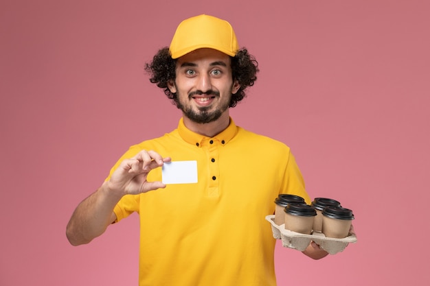 Курьер-мужчина в желтой форме с коричневыми кофейными чашками и белой карточкой на розовой стене, вид спереди