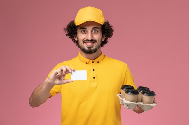 Front view male courier in yellow uniform holding brown delivery coffee cups and white card on the pink wall