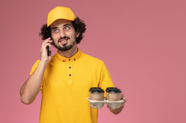 Front view male courier in yellow uniform holding brown delivery coffee cups with phone on the pink wall