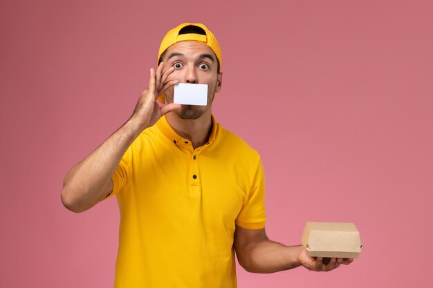 Front view male courier in yellow uniform holding card and little food package on pink background.