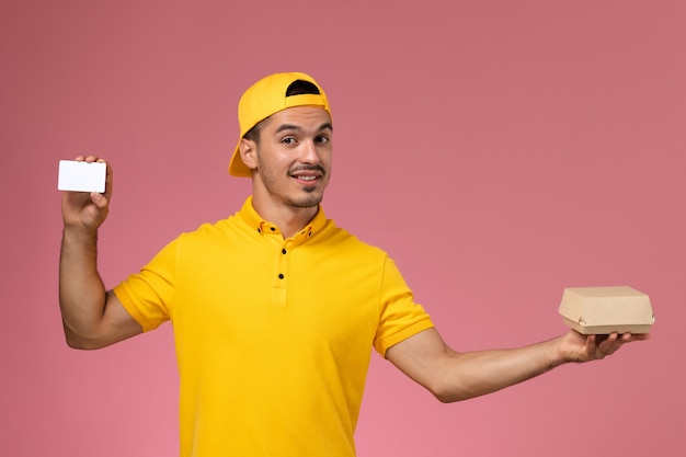 Курьер мужского пола вид спереди в желтой форме держа карточку и небольшой пакет еды на розовом фоне.