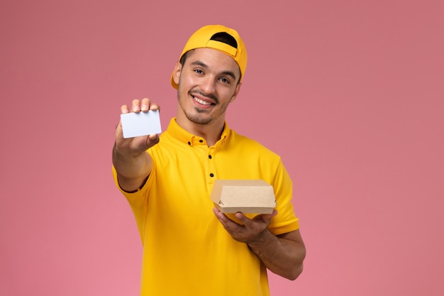 Front view male courier in yellow uniform holding card and little food package on pink background.