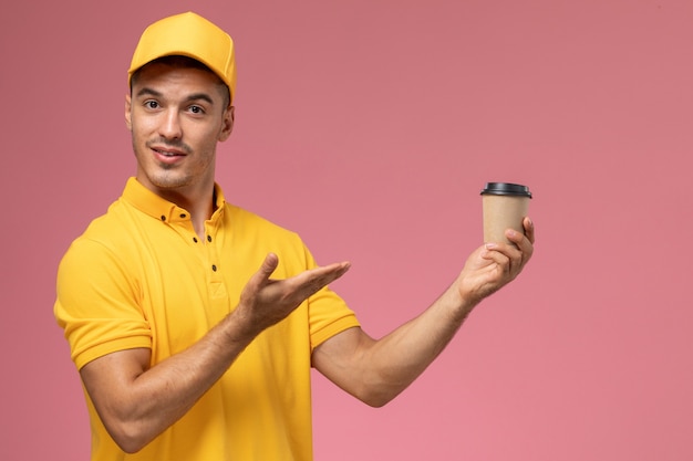 Foto gratuita corriere maschio di vista frontale in uniforme gialla che tiene tazza di consegna del caffè sui precedenti rosa