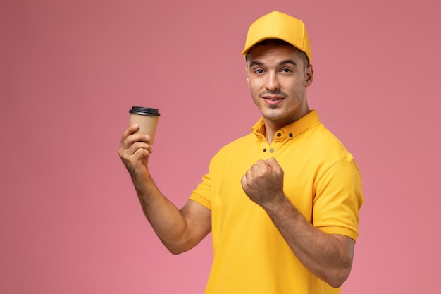 Front view male courier in yellow uniform holding coffee delivery cup on pink desk  