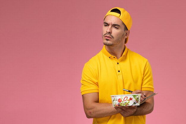 Front view male courier in yellow uniform holding delivery bowl along with notepad on pink background.