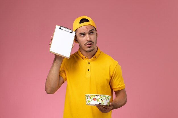 Front view male courier in yellow uniform holding delivery bowl along with notepad thinking on light pink background.