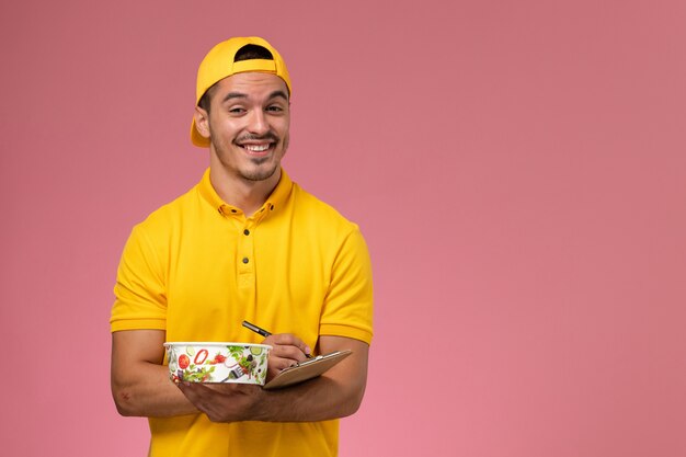 Front view male courier in yellow uniform holding delivery bowl along with notepad writing notes on the pink desk.