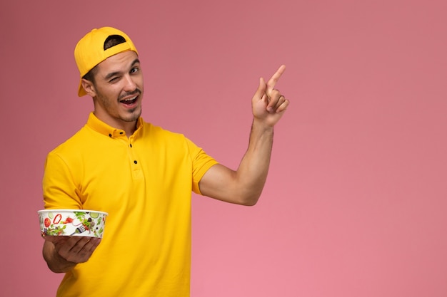 Front view male courier in yellow uniform holding delivery bowls on the light-pink background.