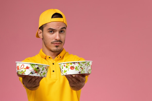 Front view male courier in yellow uniform holding delivery bowls on the pink background.