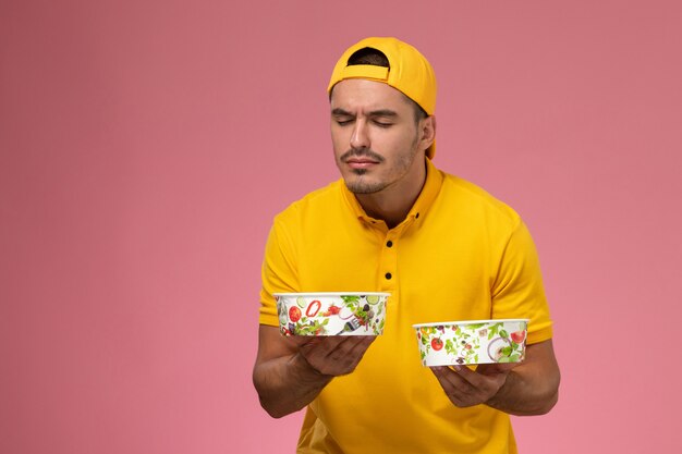 Front view male courier in yellow uniform holding delivery bowls smelling on the pink background.