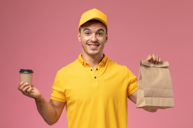 ピンクの机の上の配達のコーヒーカップと食品パッケージを保持している黄色の制服を着た正面男性宅配便