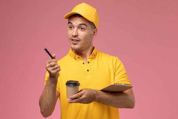 Курьер-мужчина, вид спереди в желтой форме, держит чашку кофе и блокнот, записывает заметки на розовом столе