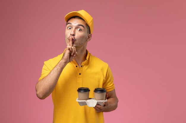 Курьер-мужчина в желтой униформе с доставкой кофе, просящий молчать на светло-розовом фоне, вид спереди