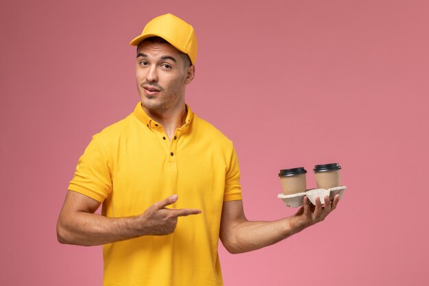 Front view male courier in yellow uniform holding delivery coffee cups on light-pink background  