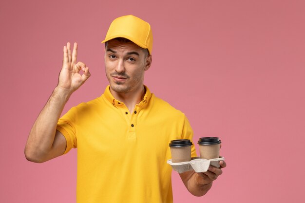 Front view male courier in yellow uniform holding delivery coffee cups on light-pink background  