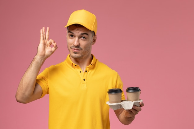 밝은 분홍색 배경에 배달 커피 컵을 들고 노란색 제복을 입은 전면보기 남성 택배