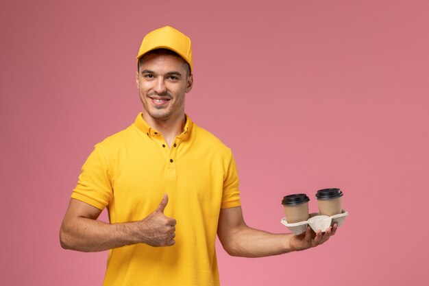 Front view male courier in yellow uniform holding delivery coffee cups on the light-pink background  