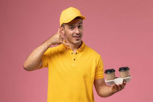 Front view male courier in yellow uniform holding delivery coffee cups phone call posing on light-pink background  