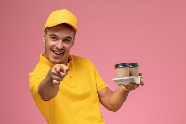 Курьер-мужчина в желтой униформе, вид спереди, радуется доставке кофейных чашек на розовом столе
