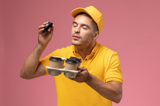 Курьер-мужчина в желтой форме, вид спереди, держит кофейные чашки и нюхает их на розовом столе