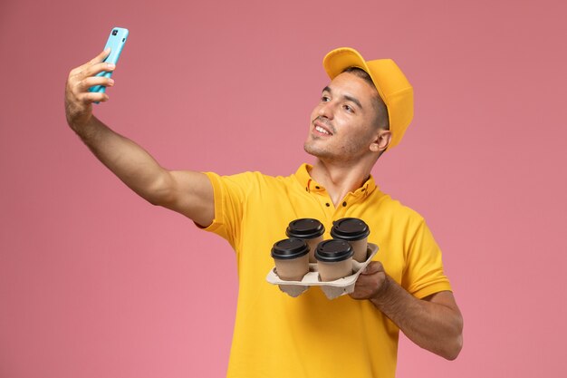 Front view male courier in yellow uniform holding delivery coffee cups taking photo with them on pink desk 