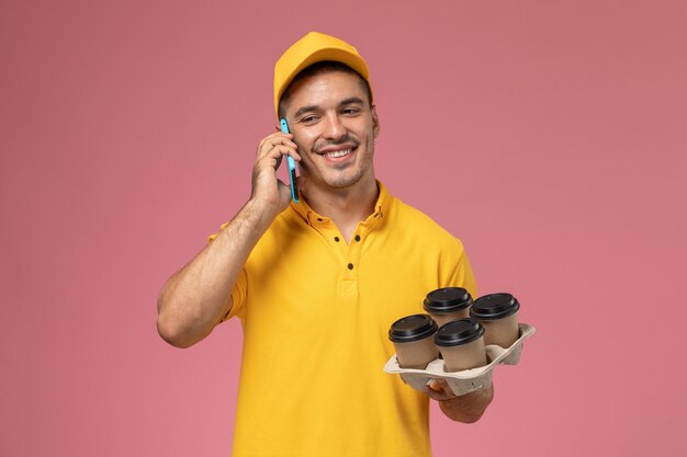 Front view male courier in yellow uniform holding delivery coffee cups and talking on the phone with smile on pink desk 