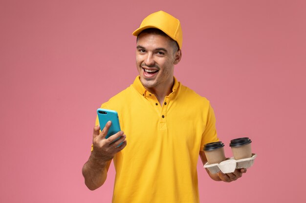 Free photo front view male courier in yellow uniform holding delivery coffee cups using his phone laughing on pink background