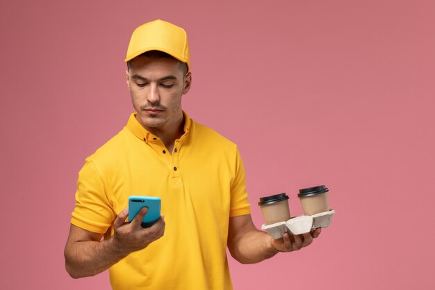 Front view male courier in yellow uniform holding delivery coffee cups using his phone on pink background  