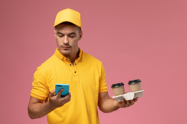 ピンクの背景に彼の電話を使用して配信のコーヒーカップを保持している黄色の制服を着た正面男性宅配便