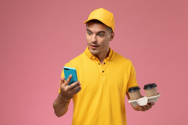 Front view male courier in yellow uniform holding delivery coffee cups using his phone on pink desk  