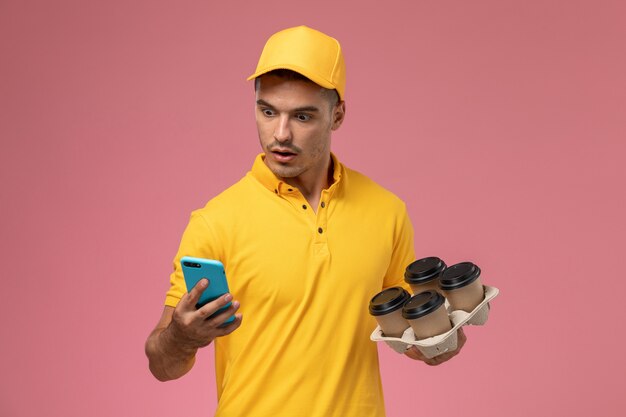 ピンクの背景に彼の電話を使用しながら配信のコーヒーカップを保持している黄色の制服を着た正面男性宅配便