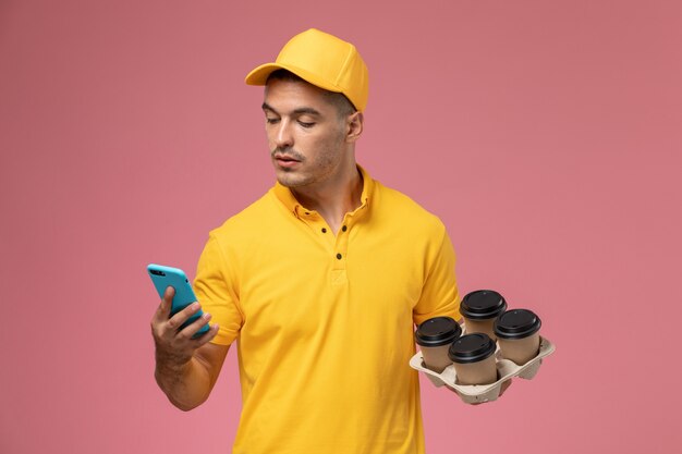 Front view male courier in yellow uniform holding delivery coffee cups while using his phone on pink desk 