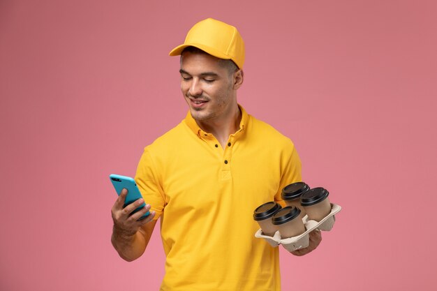 Free photo front view male courier in yellow uniform holding delivery coffee cups while using his phone on the pink desk