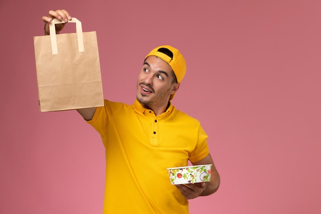 Front view male courier in yellow uniform holding delivery food package bowl on pink background.