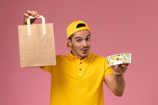 Front view male courier in yellow uniform holding delivery food package bowl on pink background.