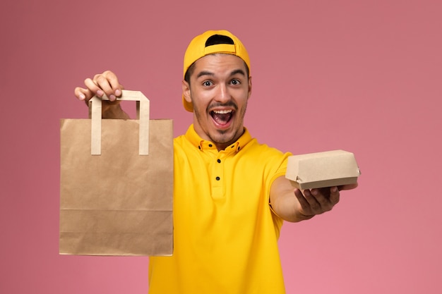 Front view male courier in yellow uniform holding delivery food packages cheering on the light pink desk.