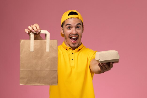 淡いピンクの机の上で応援する配達食品パッケージを保持している黄色の制服を着た正面図の男性宅配便。