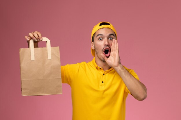 Front view male courier in yellow uniform holding delivery paper package calling out on light pink background.